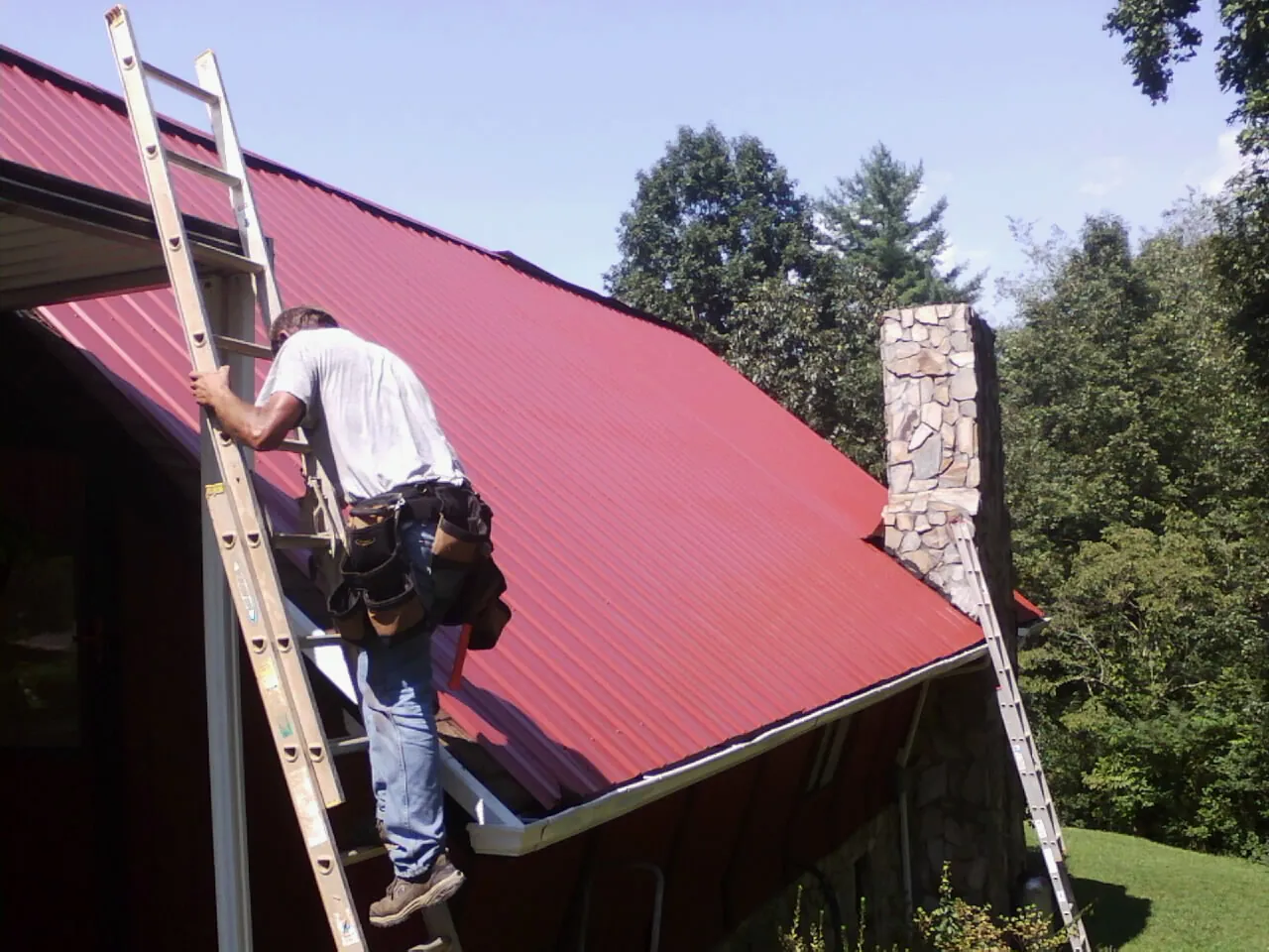 Roofer on a ladder beside a red metal roof and stone chimney — Soffit & Fascia Repair by Roofing Brothers in Osakis